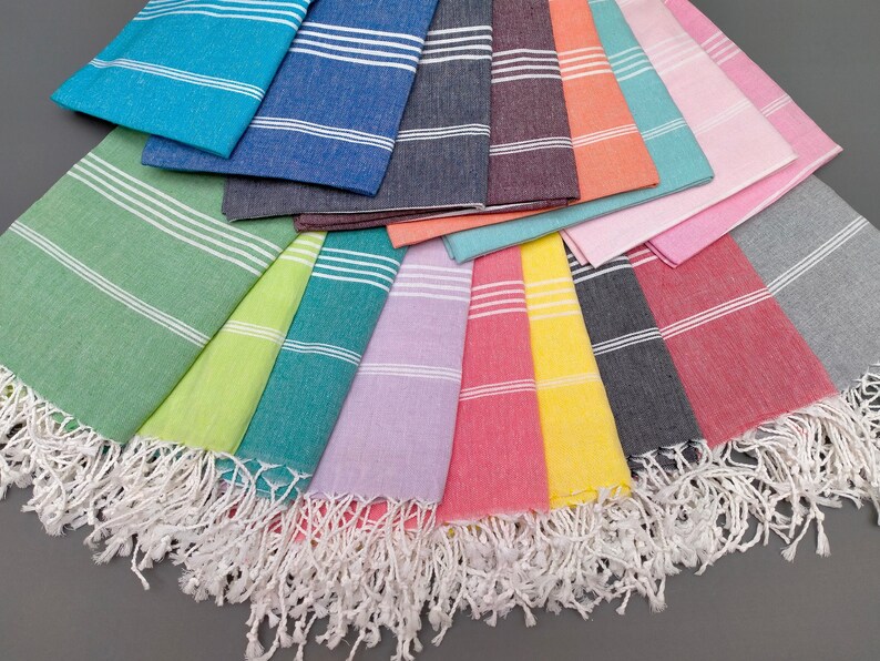May include: A collection of 14 Turkish towels in various colors, including green, yellow, teal, lavender, pink, yellow, gray, and red. The towels have white stripes and white fringe.