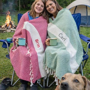May include: Two people wrapped in blankets, sitting in blue camping chairs near a campfire. One blanket is pink with a white stripe and the name "Brin". The other is green with a white stripe and the name "Cari". A dog is in the foreground.