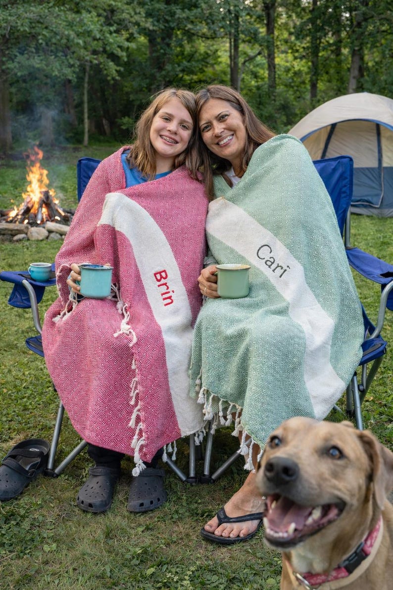 May include: Two people wrapped in blankets, sitting in blue camping chairs near a campfire. One blanket is pink with a white stripe and the name "Brin". The other is green with a white stripe and the name "Cari". A dog is in the foreground.