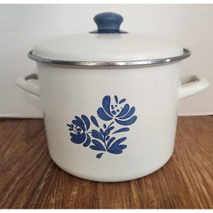 May include: White enamel pot with a blue floral design and a matching lid. The pot has two side handles and a blue knob on the lid. A silver rim accents the top of the pot. The pot is sitting on a wooden surface.