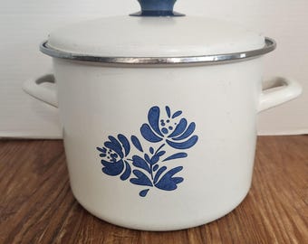 May include: White enamel pot with a blue floral design and a matching lid. The pot has two side handles and a blue knob on the lid. A silver rim accents the top of the pot. The pot is sitting on a wooden surface.