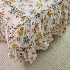 May include: White floral bed skirt with a ruffled edge. The fabric features a pattern of yellow, pink, and blue flowers. The bed skirt has a green trim along the ruffled edge, adding a decorative touch to the bedding.