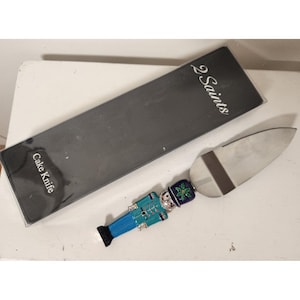May include: A cake knife with a stainless steel blade and a decorative handle. The handle is designed as a toy soldier in blue, teal, and silver, with a black base. The packaging reads "2 Saints" and "Cake Knife."
