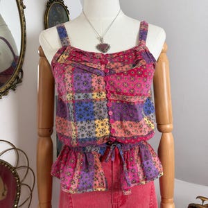 May include: A sleeveless button-down top featuring a multi-colored patchwork pattern in shades of pink, purple, orange, and blue. The garment has a gathered waist with a tie detail, a square neckline, and a peplum hem, displayed on a mannequin.