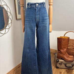 May include: Vintage blue denim flared jeans. The high-waisted pants have a button closure and a wide leg. The jeans are displayed on a mannequin. Brown leather boots and a wicker basket are in the background.