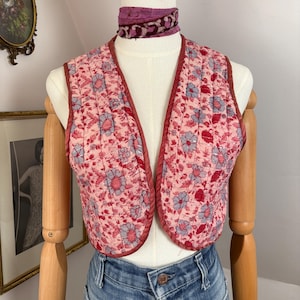 Könnte beinhalten: Eine gesteppte Weste mit einem Blumenmuster in Rosa, Rot und Blau. Die Weste hat eine bordeauxfarbene Paspelierung und wird auf einer Schaufensterpuppe präsentiert. Ein gemusterter Schal liegt um den Hals der Schaufensterpuppe.