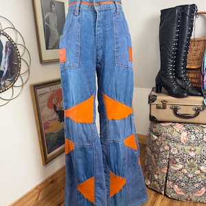 Op de afbeelding: Blauwe spijkerbroek met hoge taille en wijde pijpen, voorzien van feloranje driehoekige stoffen patches op de voorpanden. De broek heeft een knoopsluiting, riemlussen en twee voorzakken, afgewerkt met oranje stiksels langs de tailleband.