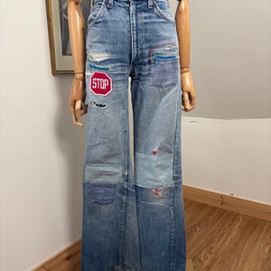 Puede incluir: Vaqueros de mezclilla azul claro con parches y una cinturilla a rayas rojas y blancas. Los vaqueros presentan un parche de señal de stop roja y varios otros parches. Los vaqueros se exhiben en un maniquí.