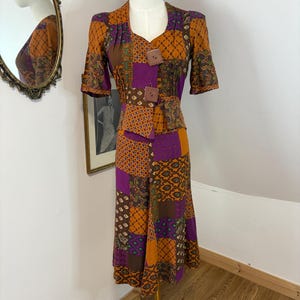 May include: A vintage patchwork dress in shades of orange, purple, and brown, displayed on a mannequin. The dress features short sleeves, a button-up bodice, and a knee-length skirt. The fabric has a variety of geometric and floral patterns.