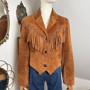 May include: A tan suede jacket with fringing across the chest and shoulders. The jacket features a classic collar, two front pockets, and three black buttons. The jacket is displayed on a mannequin with blue jeans.