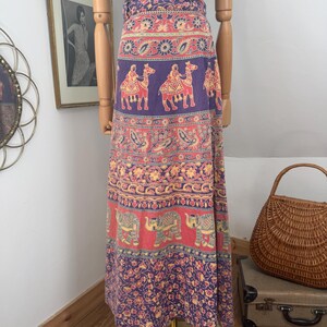 May include: A floor-length wrap skirt featuring a vibrant multi-colored print with horizontal bands of camels, elephants, and floral patterns in shades of purple, red, and yellow, worn with a bright blue sleeveless top with a white floral design.