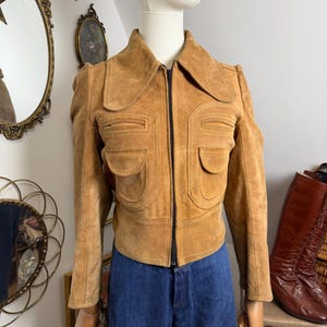 Op de afbeelding: Een beige suède jas met een grote kraag, een centrale zwarte rits en twee voorzakken met flappen. De jas heeft lange mouwen en een gestructureerde pasvorm, getoond op een etalagepop die een blauwe spijkerbroek draagt.