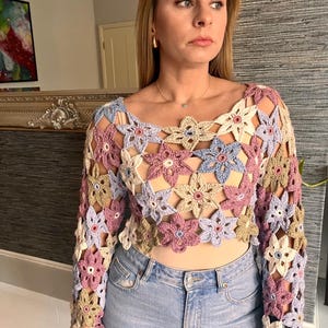 May include: A crocheted top with a floral design in shades of pink, blue, and beige. The top has long sleeves and a cropped length.