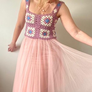 May include: A handmade crochet top in a square pattern with shades of pink, white, and blue. The top is paired with a long, flowing, light pink tulle skirt. The outfit is a mix of textures and colors.