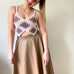 May include: A cream-colored crochet crop top with a floral pattern in shades of purple and blue. The top has thin straps and a V-neck design. The model is wearing a tan-colored skirt.