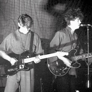 May include: Two people playing electric guitars on a stage. The person on the left is wearing a grey sweatshirt and playing a black electric guitar. The person on the right is wearing a light-coloured shirt and playing a brown electric guitar.
