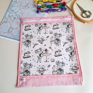 Könnte beinhalten: Eine rechteckige Stofftasche mit weißem Reißverschluss, die ein wiederkehrendes Muster aus nähenden Zeichentrickfiguren, Schneiderpuppen und Nadelkissen auf weißem Grund mit kleinen rosa Herzen zeigt und mit einem rosa Stoffrand versehen ist.