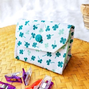 May include: A small, white fabric pouch with a flap closure, adorned with a pattern of green shamrocks. The pouch is secured with a turquoise button. Sewing clips in purple and orange are scattered in the foreground.