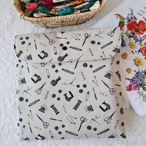 Op de afbeelding: Een beige naaigereedschapsetui met een zwart patroon van naaigerei, waaronder scharen, naalden en garenklosjes. De etui sluit met een zwarte knoop. Een mand met kleurrijk borduurgaren en een bloemenborduurwerk zijn op de achtergrond te zien.