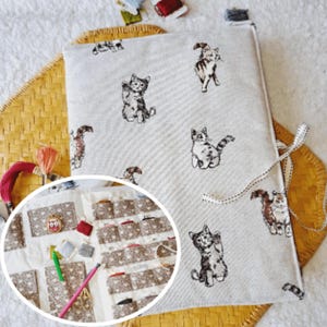 Peut inclure: Un organisateur de couture en tissu crème avec un motif répété de chats illustrés. L'organisateur a une fermeture à ruban et une incrustation circulaire montrant des poches pour les fils, les aiguilles et autres fournitures de couture. Le fond est une surface tissée brun clair.