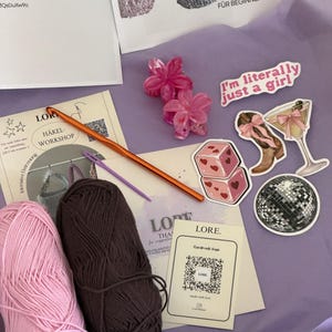 May include: A crochet kit containing two skeins of yarn in light pink and dark brown, an orange crochet hook, a purple plastic needle, two pink flower hair clips, and four stickers featuring dice, a cowboy boot, a martini, and a disco ball.