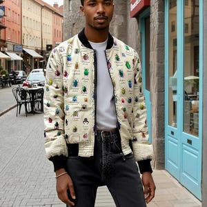 Puede incluir: Una chaqueta bomber blanca con un estampado de escarabajos coloridos. La chaqueta tiene una cremallera negra y puños y cintura de canalé negros. El modelo lleva pantalones vaqueros negros.