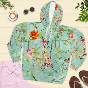 May include: A hooded sweatshirt with a floral pattern in shades of orange, pink, and green on a light teal background. The design includes butterflies and vines. The sweatshirt is paired with pink sweatpants and brown sandals.