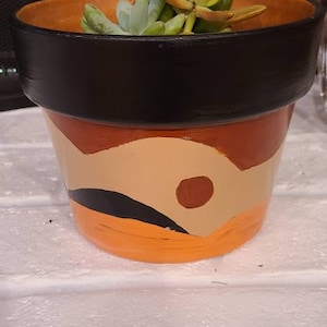 May include: A terracotta planter with a black rim and painted orange, brown and beige abstract design. The planter is filled with green succulents.