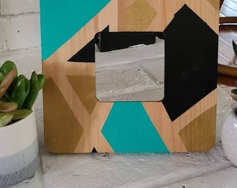 Op de afbeelding: Een houten fotolijst met een geometrisch ontwerp in teal, goud en zwart. De lijst heeft een vierkante opening in het midden.