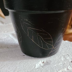 Puede incluir: Maceta de terracota negra con diseño de hojas doradas. La maceta está vacía y se encuentra sobre una superficie blanca.
