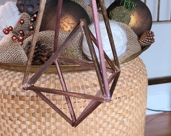 May include: A wooden geometric ornament in a brown and pink color scheme hangs from a wicker basket. The ornament is a pyramid shape with a square base. The basket is filled with burlap and natural elements.