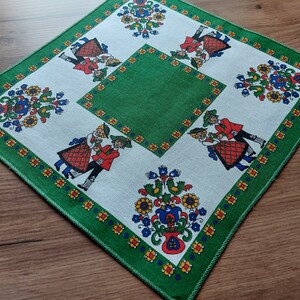 Può includere: Una tovaglia verde e bianca con un design tradizionale popolare. La tovaglia presenta un motivo ripetitivo di fiori, foglie e persone in abiti tradizionali. La tovaglia è rifinita con un bordo verde.