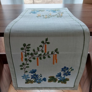 May include: White table runner with an embroidered floral design. The design features green leaves, orange stems, and blue flowers. The runner has a white background and a white border.