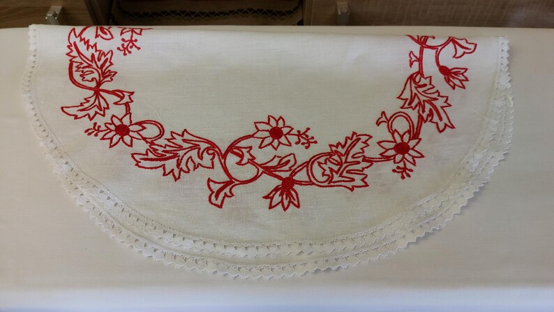 May include: White round tablecloth with a red embroidered floral design and a white crocheted lace trim.
