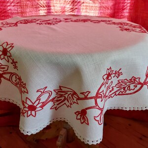 May include: White linen tablecloth with a red embroidered floral design. The tablecloth has a scalloped edge and is laid on a wooden table.