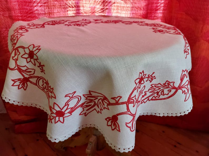 May include: White linen tablecloth with a red embroidered floral design. The tablecloth has a scalloped edge and is laid on a wooden table.