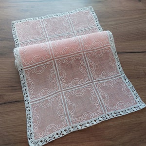 Puede incluir: Un delicado camino de mesa de crochet rosa y blanco con un patrón cuadrado. El camino de mesa está hecho de una tela fina y ligera y tiene un delicado borde de encaje.
