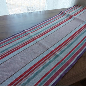 May include: A woven table runner with a striped pattern in shades of pink, red, blue, green, and purple. The runner is rectangular and appears to be made of fabric. The stripes run lengthwise, creating a visually appealing design.