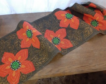 May include: A brown fabric runner with a repeating pattern of orange flowers with green centers on a dark brown background.