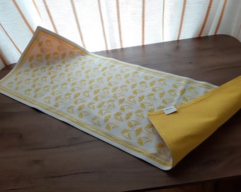 May include: A white table runner with a yellow floral pattern. The runner is folded in half and has a yellow fabric backing.