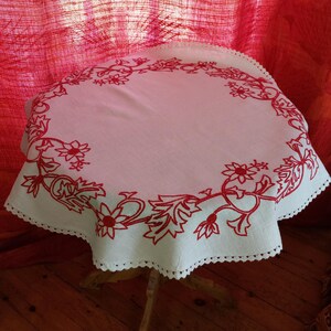 May include: White linen tablecloth with a red embroidered floral design around the edge. The tablecloth has a scalloped edge and is laid on a wooden table.