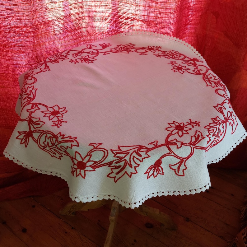 May include: White linen tablecloth with a red embroidered floral design around the edge. The tablecloth has a scalloped edge and is laid on a wooden table.