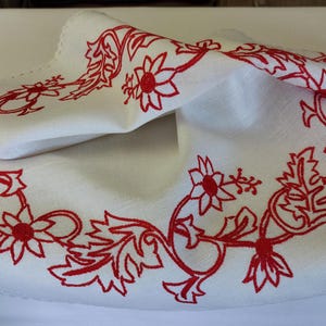 May include: White linen tablecloth with a red embroidered floral design around the edge. The tablecloth has a scalloped edge.