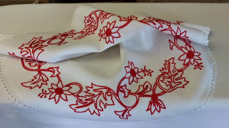 May include: White linen tablecloth with a red embroidered floral design around the edge. The tablecloth has a scalloped edge.