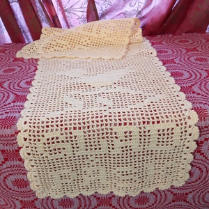 May include: A crocheted table runner in a cream color with a scalloped edge. The runner is laid out on a red and white patterned fabric.