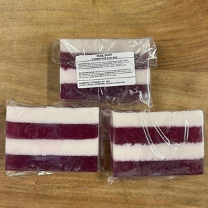 May include: Three Cornish Pixie Soap Bars, each with a layered design of white and deep purple. The bars are individually wrapped in clear plastic. The top bar has a label that reads "Pixie Dust Cornish Pixie Soap Bar".