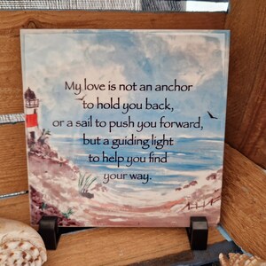 May include: Square ceramic tile with a coastal scene featuring a lighthouse, ocean, and sky. The tile has the text "My love is not an anchor to hold you back...but a guiding light to help you find your way."