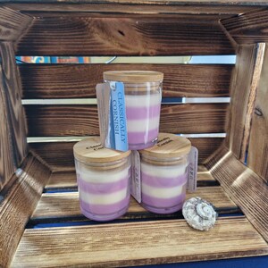 May include: Three glass jar candles with wooden lids, layered with white and lilac wax. Each candle has a tag that reads "Classically Cornish". A small seashell sits nearby, all inside a wooden crate.