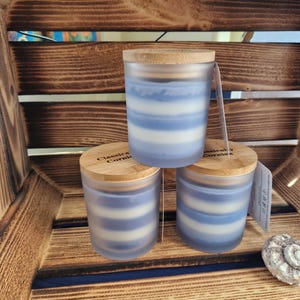 May include: Three cylindrical candles with blue and white horizontal stripes, topped with wooden lids. The candles are in frosted glass jars, and the lids have text that reads "Classics Cornwall". The candles are displayed in a wooden crate.