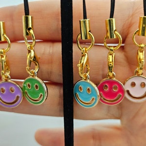 May include: Four gold charm necklaces with different colored smiley faces. The charms are attached to black cords. The smiley faces are purple, green, blue, and pink.